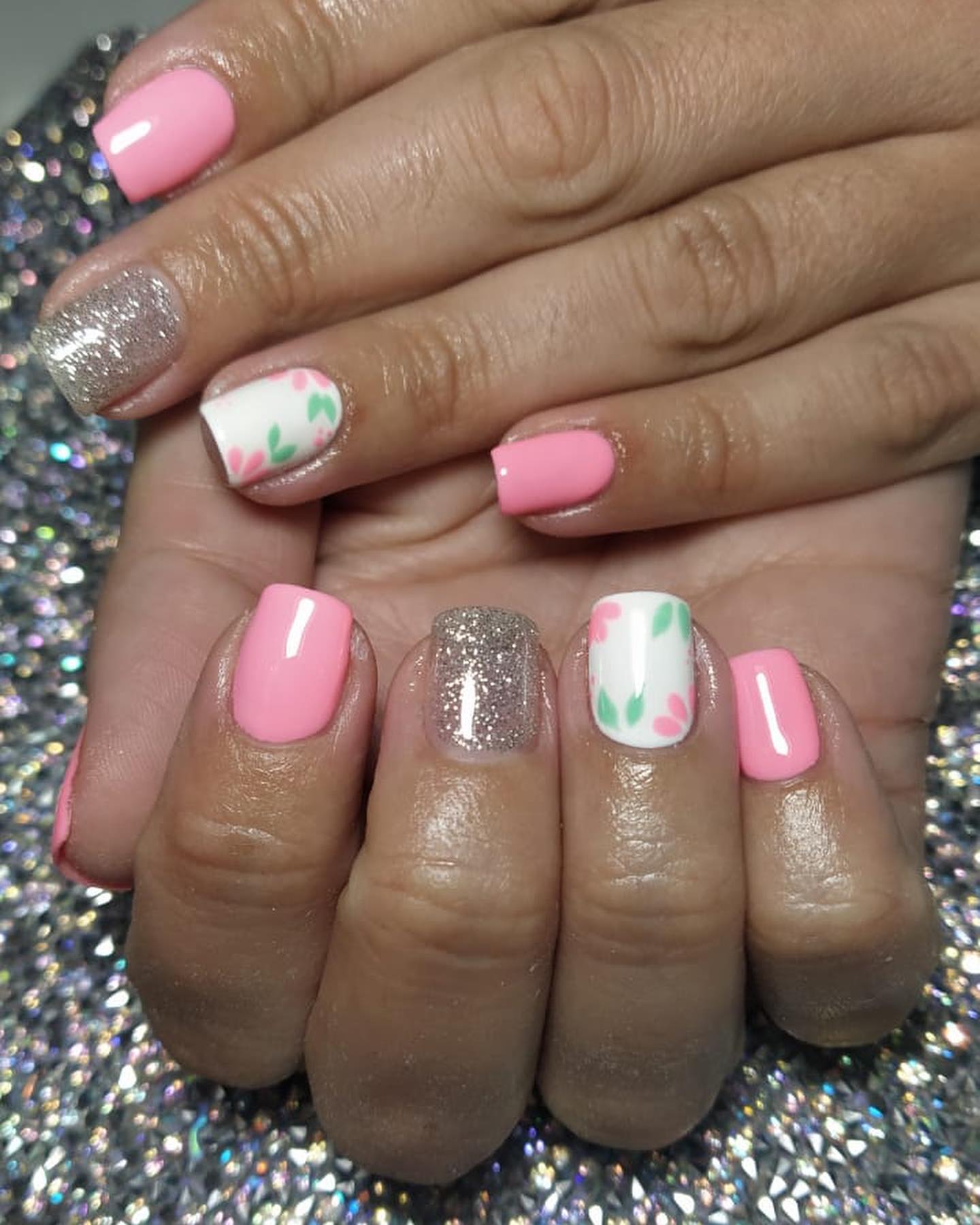 Uñas en semipermanente rosa, una con glitter plateado y otra blanca con flores rosas y hojas verdes pintadas a mano