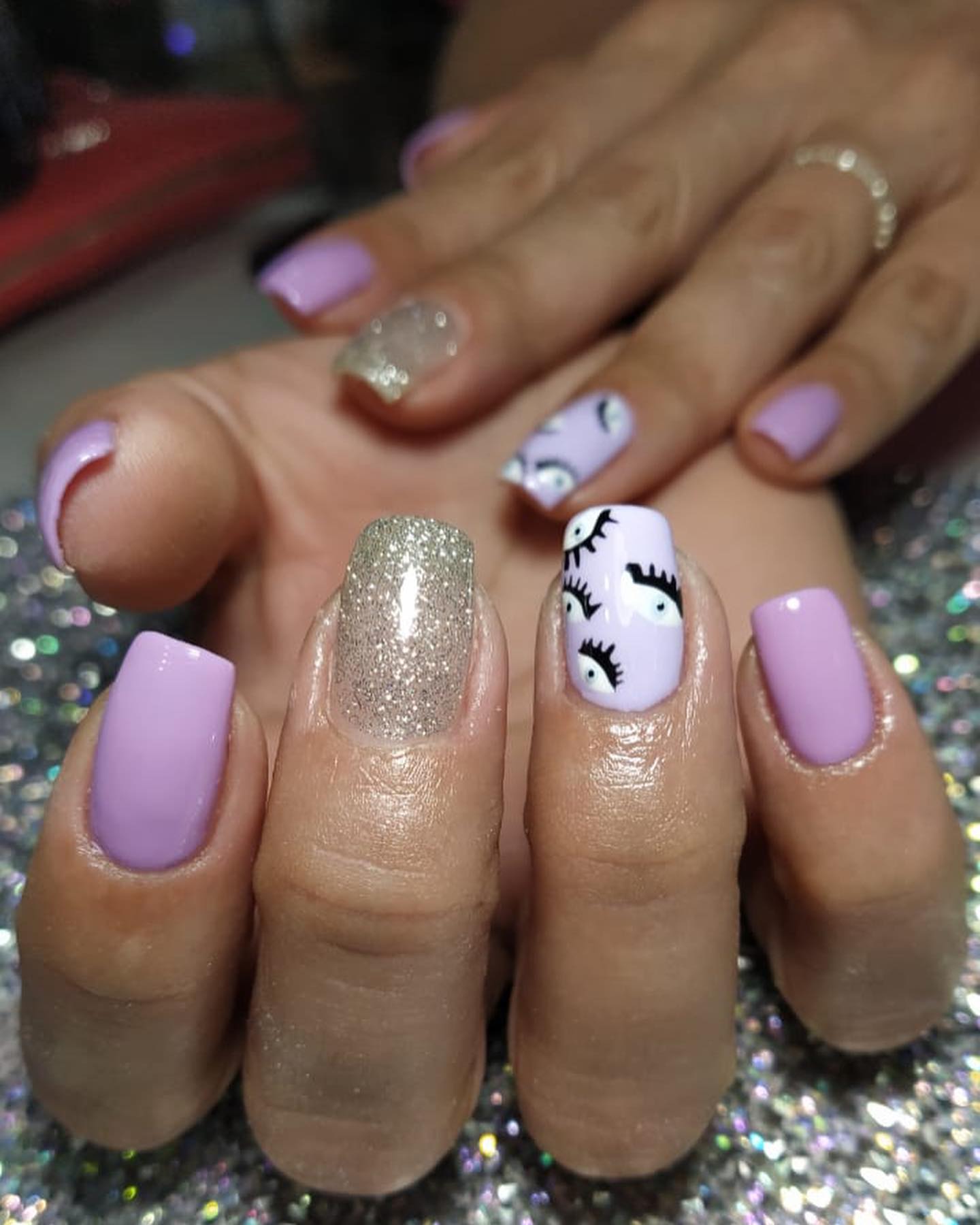 Uñas cortas en semipermanente lila, una uña con glitter plateado y ojitos pintados a mano, sobre el fondo de glitter de GlamB